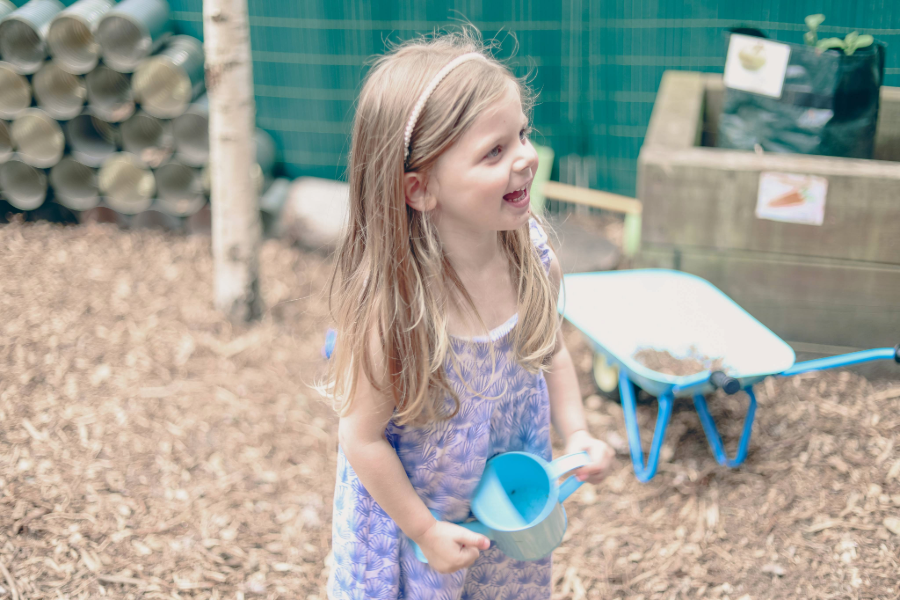 Young girl smiling, holding a watering can in an outdoor play area with wood chips.