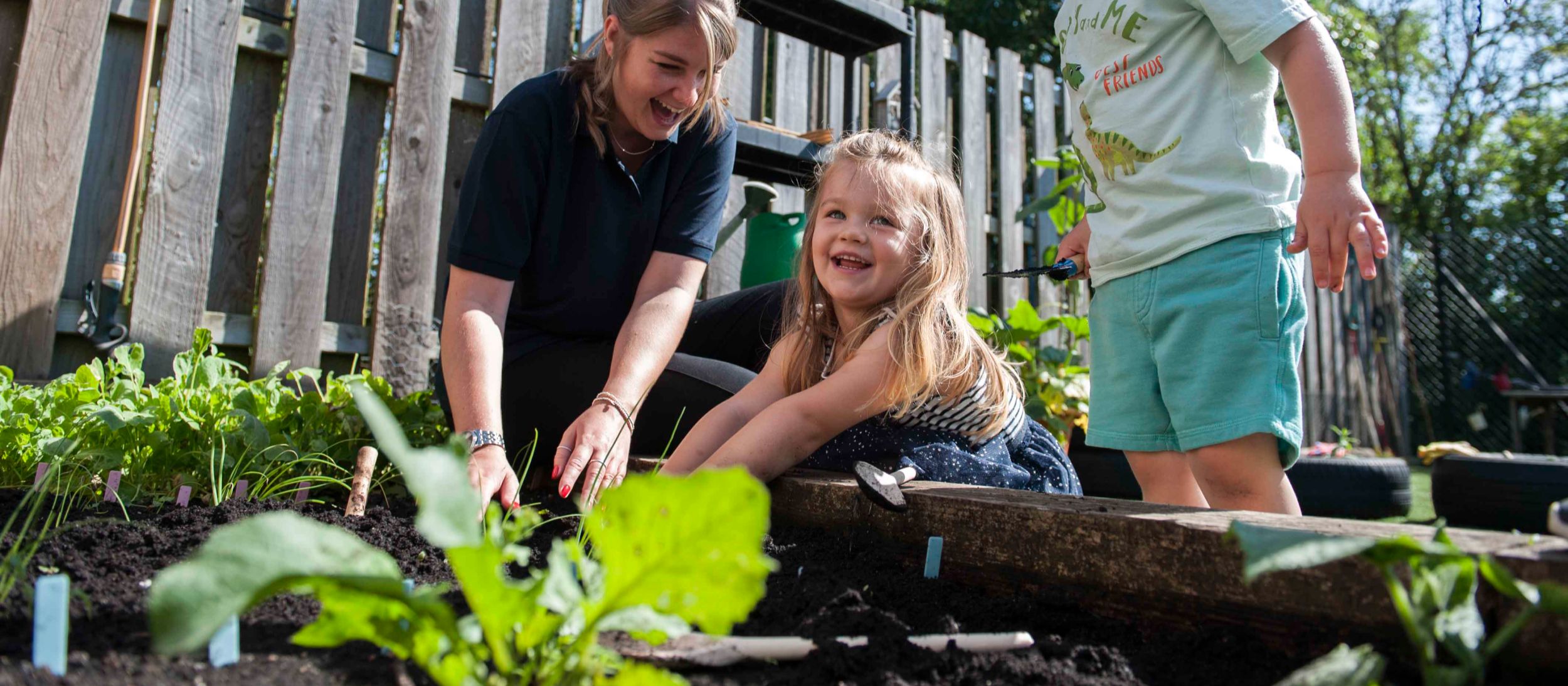 A teacher and two children are happily planting seedlings in an outdoor raised garden bed at the nursery.