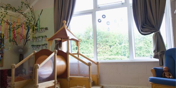 An indoor play area with a wooden climbing structure, a window overlooking a garden, and a chair with a doll.