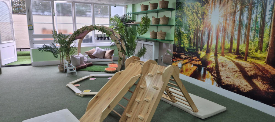 Indoor play space with wooden climbing structures, nature-themed mural, and a quiet corner.