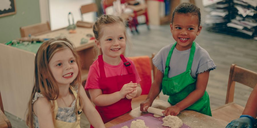 Three happy children wearing aprons, kneading dough with rolling pins at a table.