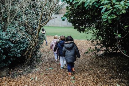 A group of children are walking single-file along a leafy path in a garden at the nursery.