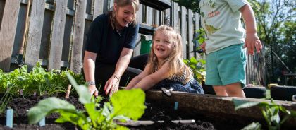 A teacher and two children are happily planting seedlings in an outdoor raised garden bed at the nursery.
