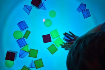 Close-up of hands playing with colored translucent geometric blocks on a bright blue light table.