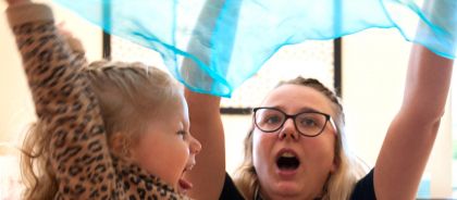 A teacher and a child raise a sheer blue scarf over their heads, their mouths open in a joyful expression.